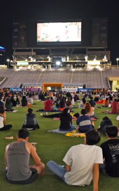 students sitting on the football field at night watching a movie on the video board