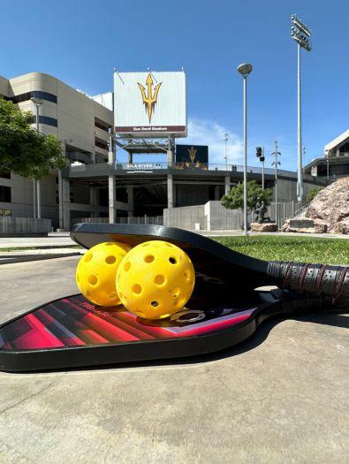 Yellow whiffle-style balls stacked between two small flat paddles balance in front of Sun Devil Stadium