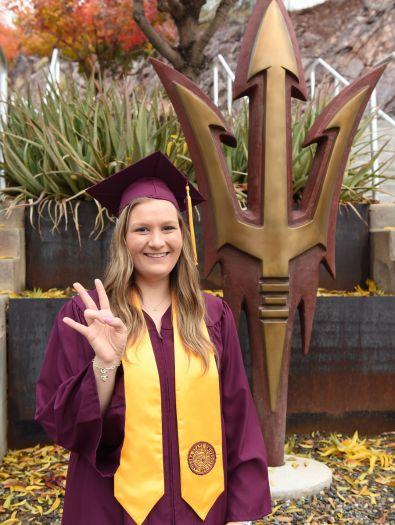 female student in cap and gown smiling at the camera with a pitchfork statue in the background